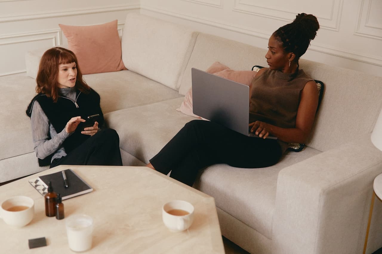 Two women sitting down in a living room chatting with a laptop and cellphone in hand. 